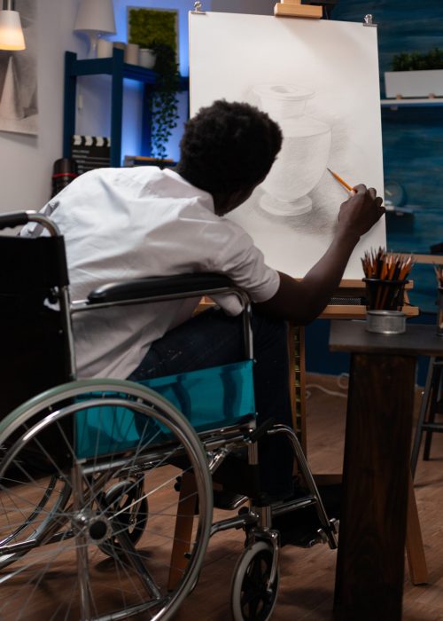 Art student in wheelchair sketching vase ilustration working at shadow using professional graphic tools during creative class. Young man illustrator drawing on canvas having inspiration in studio