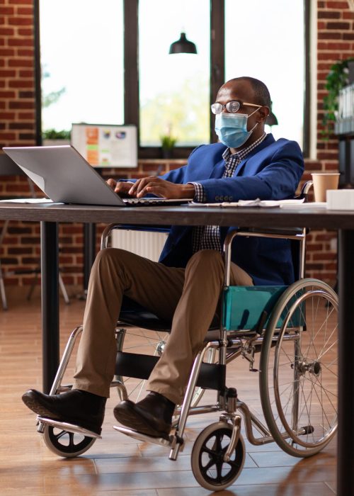 Employee with disability wearing face mask and working on business project. Entrepreneur sitting in wheelchair, doing financial and marketing work during coronavirus pandemic. Man at job